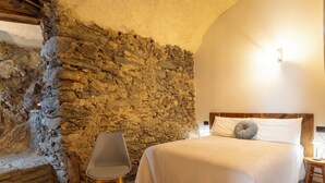 Un dormitorio con pared de piedra, cama con ropa de cama blanca, una silla gris y una mesita de noche de madera.