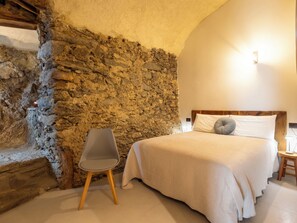 Une chambre avec un mur en pierre, un lit avec une literie blanche, une chaise grise et une table de chevet en bois.