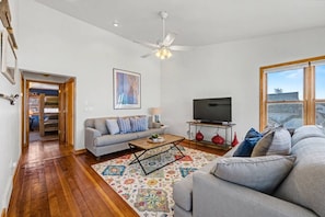 A living room with a sofa, a coffee table, a TV on a stand, a ceiling fan, and a large window. - Walkable Egg Harbor Retreat with Water Views (Egg Harbor)
