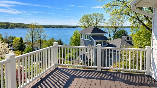 Une terrasse en bois avec une balustrade blanche, donnant sur un lac et une maison dotée d’une petite tour.