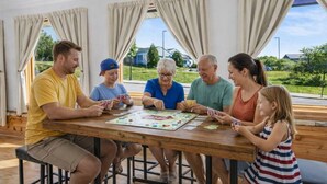Une famille joue à un jeu de société autour d’une table en bois, près de grandes fenêtres et de rideaux.
