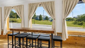 Une pièce avec une table en bois et quatre tabourets de bar noirs, de grandes fenêtres avec des rideaux beiges, et une vue sur une route et la verdure environnante.