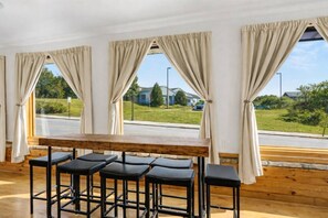 A room with a wooden table and four black bar stools, large windows with beige curtains, and a view of a road and greenery outside. - Walkable Egg Harbor Retreat with Water Views (Egg Harbor)