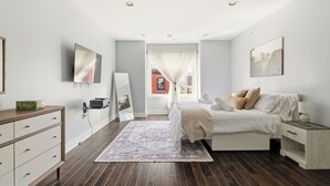 Ein Schlafzimmer mit Bett, Nachttisch, Kommode und einem großen Fenster mit Vorhängen.