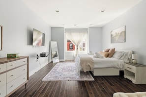 A bedroom with a bed, bedside table, dresser, and a large window with curtains.