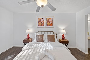Un dormitorio con una cama, dos mesitas de noche, un ventilador de techo y dos cuadros colgados en la pared.