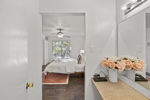 Une chambre à coucher avec un lit, un ventilateur de plafond, une fenêtre, un miroir et un vase avec des fleurs.