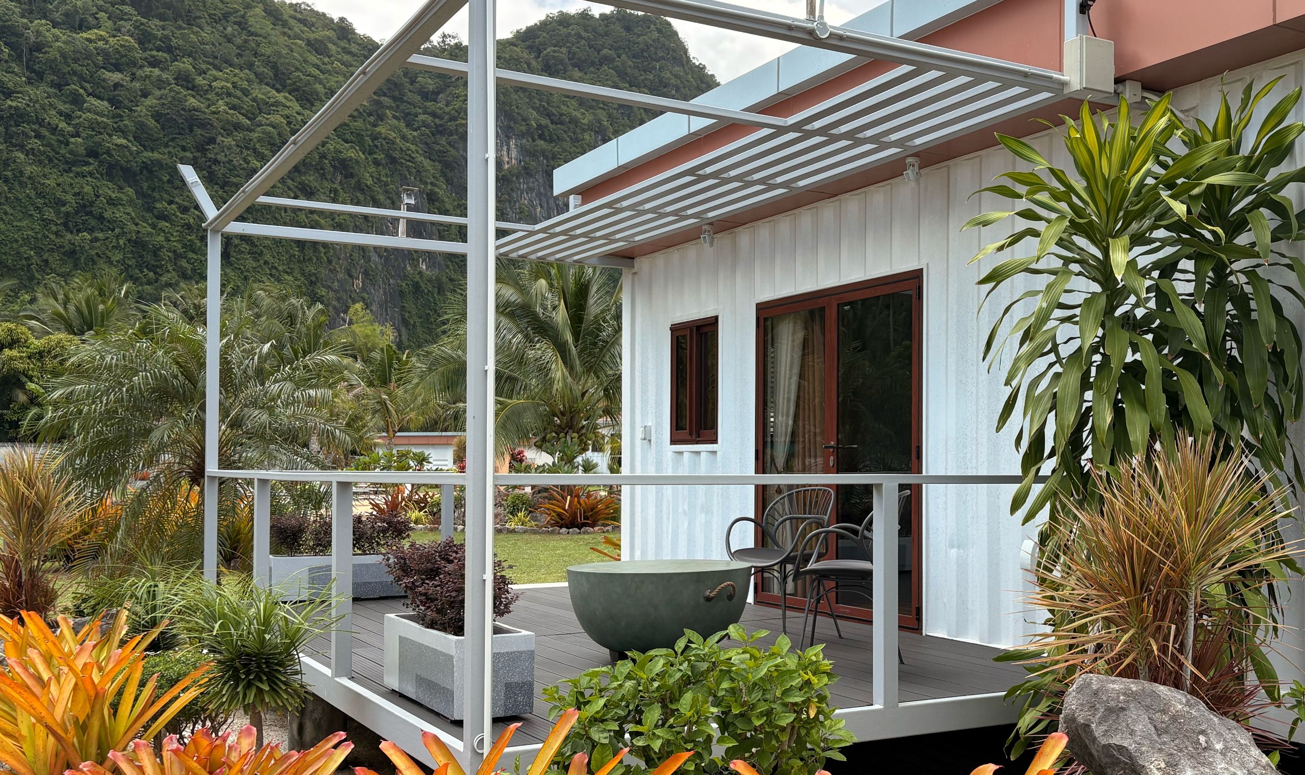 A modern house with a balcony, surrounded by tropical plants and a mountainous backdrop.