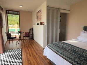 A hotel room with a bed, a desk with a chair, a view of the outdoors, and a bathroom door. - This Great Place (Don Sak)