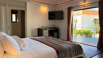 A hotel room with a bed, a desk with a flat-screen TV, a balcony with a view of greenery, and a sliding door.
