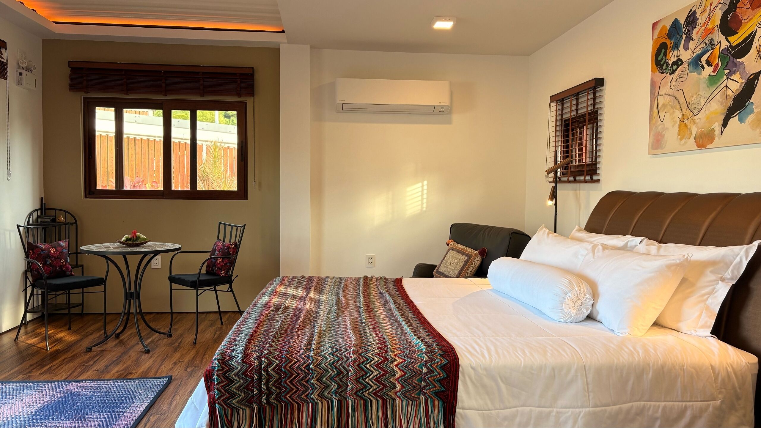 Una habitación de hotel con cama, una mesita pequeña con sillas, un equipo de aire acondicionado y una ventana con persianas.