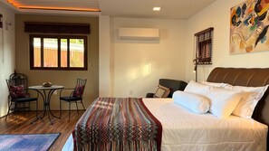 A hotel room with a bed, a small table with chairs, an air conditioning unit, and a window with blinds. - This Great Place (Don Sak)