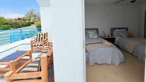 Un balcone con sedie e tavolo in legno, una camera da letto con due letti e un comodino, e vista sulla zona circostante.