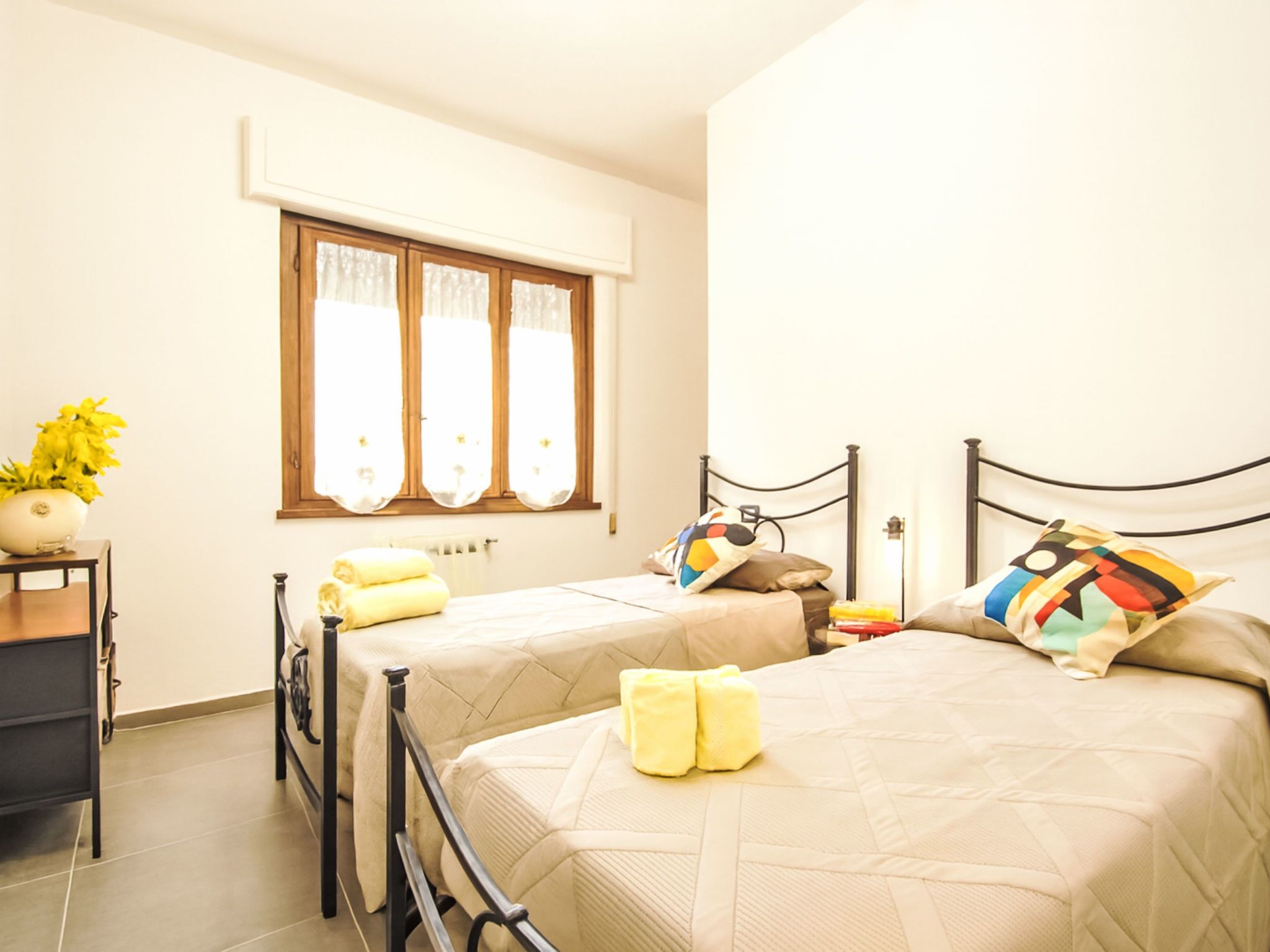 A bedroom with two beds, a window with curtains, a bedside table with a plant, and a radiator.