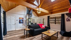 A spacious loft with a wooden ceiling, a black sofa, a wooden coffee table, and a spiral staircase. - NEW Kink Cabin w/ Private Creek (Sevierville)