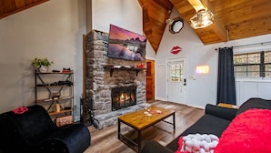 A cozy living room with a stone fireplace, a black sofa, a wooden coffee table, and a red fluffy throw. - NEW Kink Cabin w/ Private Creek (Sevierville)