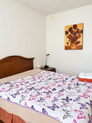 Ein Schlafzimmer mit einem hölzernen Kopfende, einem Bett mit einer zarte Schmetterlingsdecke, einem Nachttisch mit einer Lampe und einem Wandbild von Sonnenblumen.