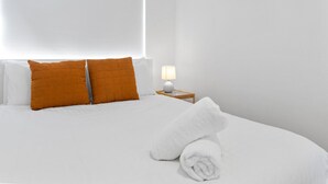 Hotellihuone, jossa on sänky, kaksi tyynyä, yöpöytä lampun kanssa ja sängyllä rullalle taitettu pyyhe.