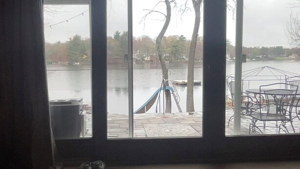 A view through a window of a riverside deck with a metal table and chairs, a blue hammock, and a ladder leaning against a tree.