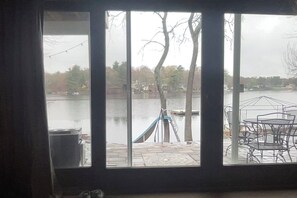 Uitzicht door een raam op een terras aan de rivier met een metalen tafel en stoelen, een blauwe hangmat en een ladder die tegen een boom leunt.