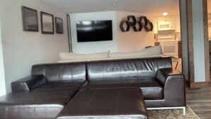 A living room with a dark leather sofa, a flat-screen TV mounted on the wall, and a kitchen area in the background.