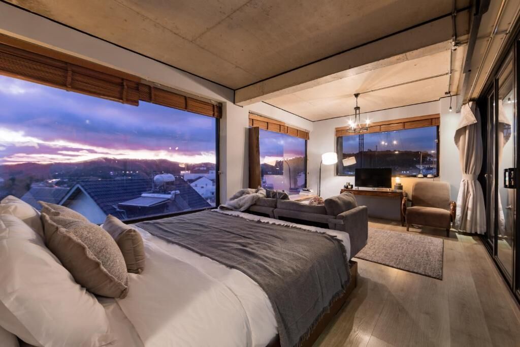 Ein modernes Schlafzimmer mit einem großen Bett, einer Couch, einem Fernseher und Blick auf eine Stadtlandschaft.