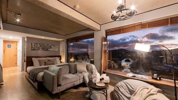 A modern bedroom with a bed, sofa, and a large window with a cityscape view.
