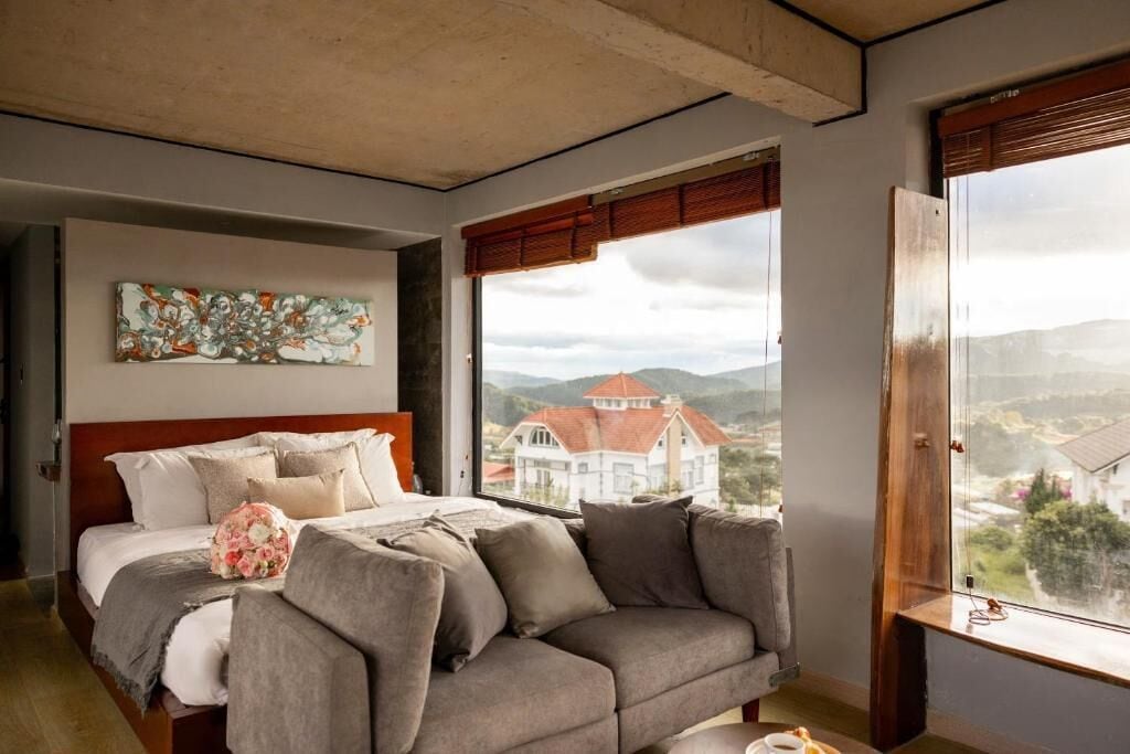 A bedroom with a bed, a gray sofa, a painting on the wall, and a window with a view of a house.