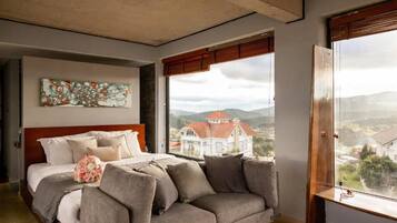 A bedroom with a bed, a gray sofa, a painting on the wall, and a window with a view of a house.
