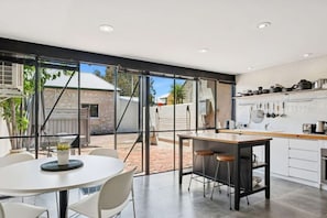 Une cuisine moderne dotée d’un îlot central, de meubles blancs et offrant une vue sur une terrasse extérieure grâce à de grandes portes vitrées.