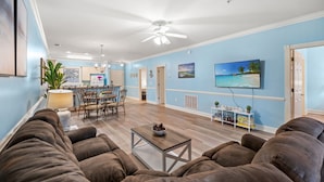Un salon avec un canapé marron, une table basse, un coin repas avec une table et des chaises, un ventilateur de plafond et un téléviseur fixé au mur.