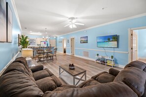 A living room with a brown sofa, a coffee table, a dining area with a table and chairs, a ceiling fan, and a TV mounted on the wall. - Ground Floor 2BR Near Golf & Beach (Myrtle Beach)