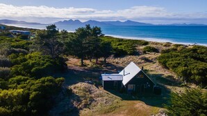 A remote cabin with a metal roof, surrounded by dense vegetation and overlooking a coastal area with mountains in the distance.