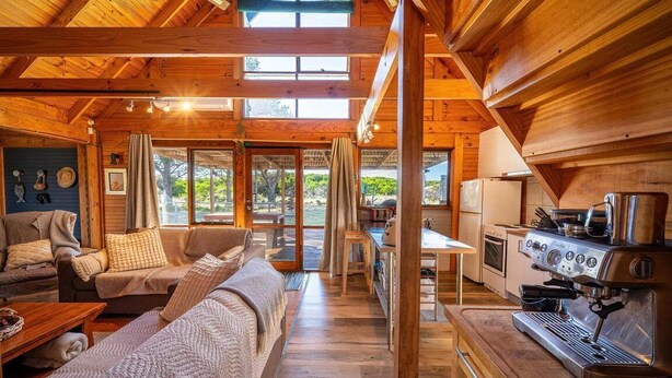 A cozy cabin interior with a living area, sofa, coffee table, and a kitchenette with a coffee machine.