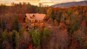 A log cabin with a prominent porch and multiple windows, surrounded by a forest with autumn foliage. - New Cabin! Family Fun - 6 BR - Smoky Mountains Views, Hot Tub, Arcades (Gatlinburg)