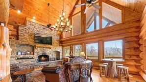 A log cabin interior with a stone fireplace, wooden furniture, and large windows. - New Cabin! Family Fun - 6 BR - Smoky Mountains Views, Hot Tub, Arcades (Gatlinburg)