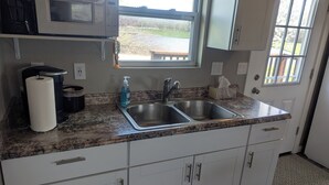 A kitchen with a granite countertop, stainless steel sink, white cabinets, a microwave, and a window with a view of a garden.