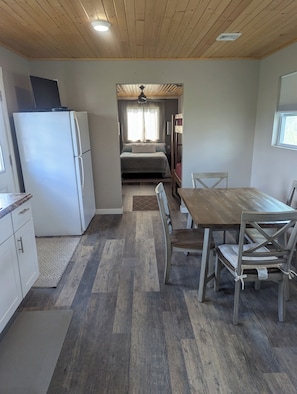 A compact kitchen with a refrigerator, a dining table with chairs, a wooden ceiling, and a bed with a window in the background.