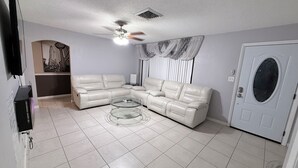 Un salon comprenant un canapé d’angle, une table basse en verre, un ventilateur de plafond, une porte avec une fenêtre et un téléviseur fixé au mur.