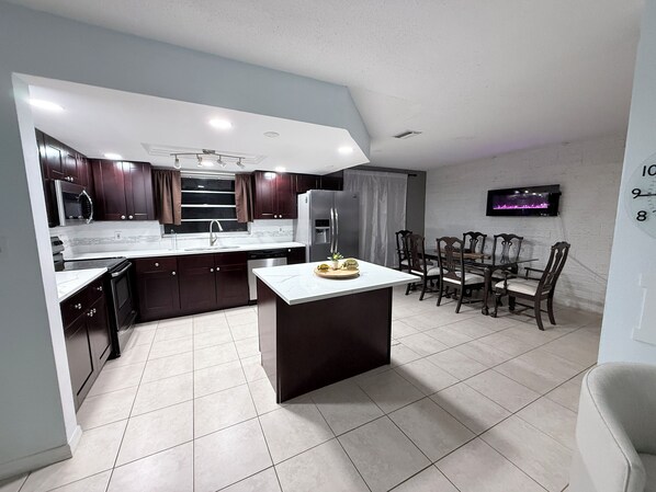 A modern kitchen with dark wood cabinets, a white countertop, and a dining area with wooden chairs and a table. - Lovely full home near Tampa, beaches, Weeki Wachee, Tarpon Springs,& Clearwater (Port Richey)