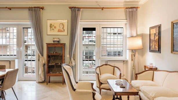 A room with a sofa, chairs, a table, a lamp, a bookshelf, and a window with a view of buildings.