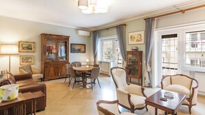 A spacious living room with a dining table, multiple chairs, a sofa, a cabinet, and a window with curtains.