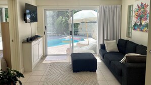 A living room with a sofa, ottoman, TV, ceiling fan, and a view of a swimming pool through glass doors. - Tranquility on Buderim - large, light family home with expansive views. (Buderim)