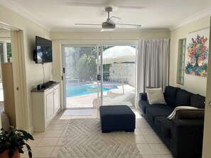 Ein Wohnzimmer mit Sofa, Hocker, Fernseher, Deckenventilator und Blick auf einen Swimmingpool durch Glastüren.