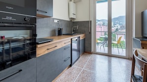 Una cocina moderna con horno empotrado, cafetera y lavavajillas. Cuenta con una encimera de madera, un fregadero y una zona de comedor con una silla. La habitación tiene un ventanal con vistas a edificios y colinas.
