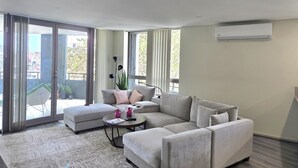 A modern living room with a sectional sofa, a small round table, and a sliding glass door leading to a balcony. - Cozy Modern Stayz | Free Parking | 1-Min Station, 5-Min Hospital | Heated Pool (Wollongong)