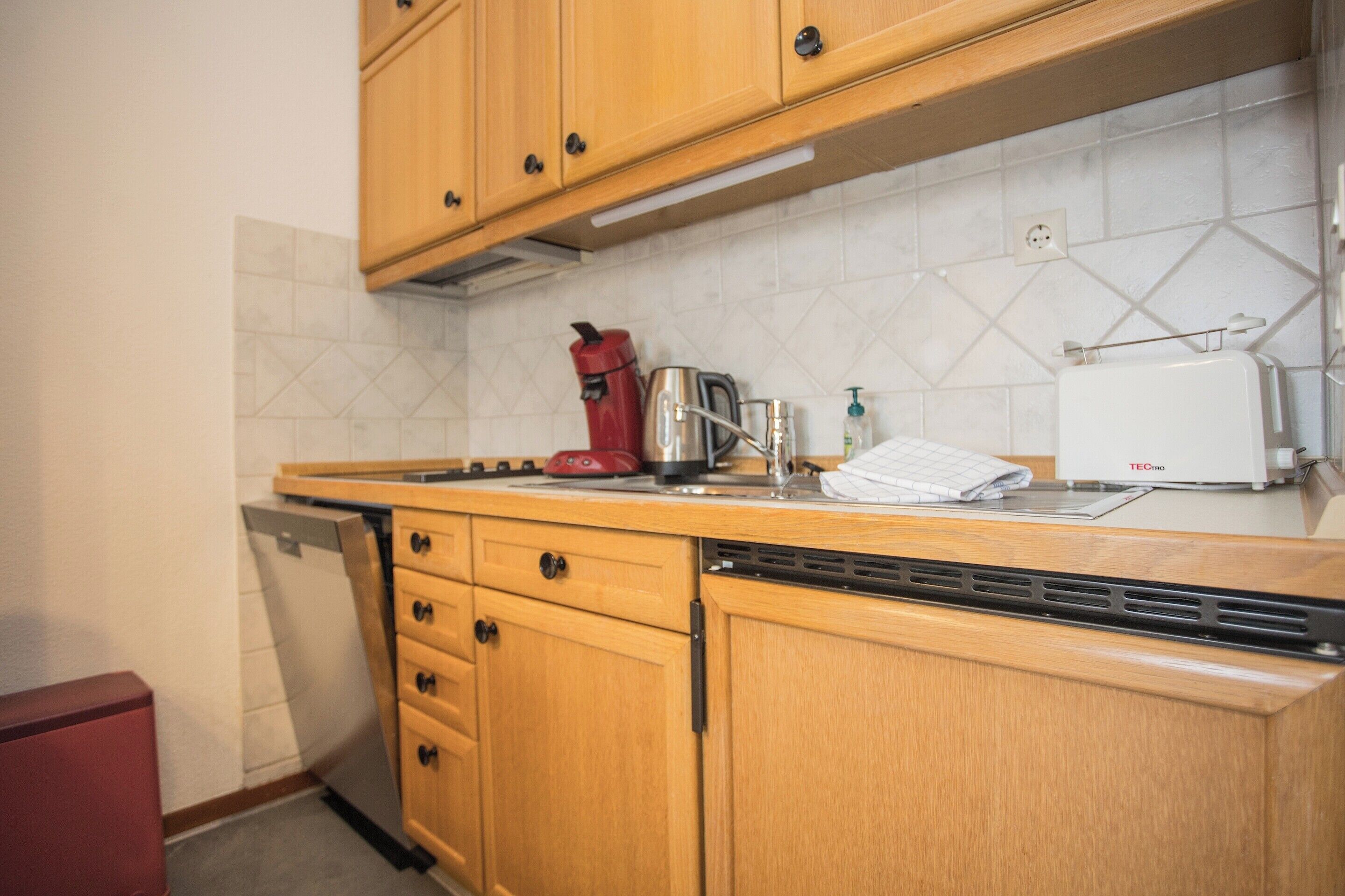 A kitchen with wooden cabinets, a dishwasher, a red kettle, a toaster, and a white appliance on the counter.