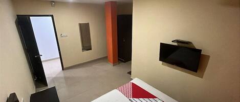 A hotel room with a bed, a TV mounted on the wall, a desk with a chair, and a door leading to another room.