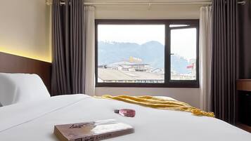 Habitación de hotel con cama, ventana con cortinas, vista a edificios y una mesita con un libro y una botella.