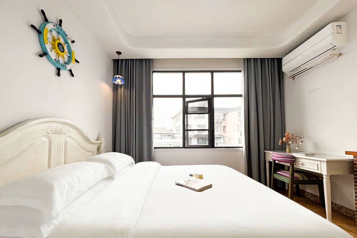 A hotel room with a bed, a desk, a window with curtains, an air conditioning unit, and a decorative wall piece.